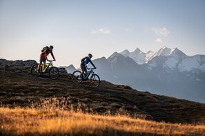 Dois ciclistas em uma trilha de montanha, com vista para montanhas cobertas de neve ao fundo.