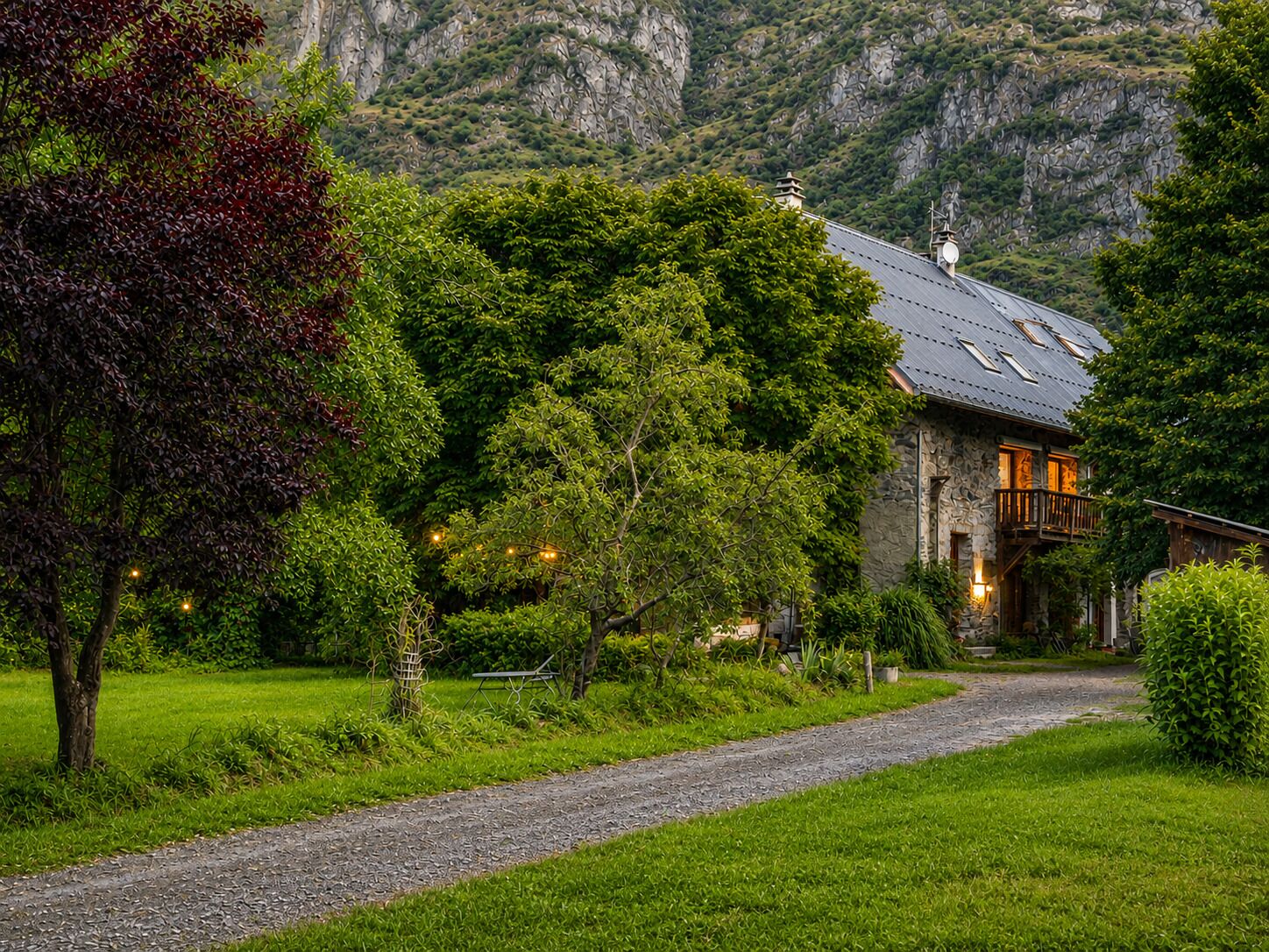 A stone house with a sloped roof, surrounded by lush greenery and a gravel path leading to the entrance.