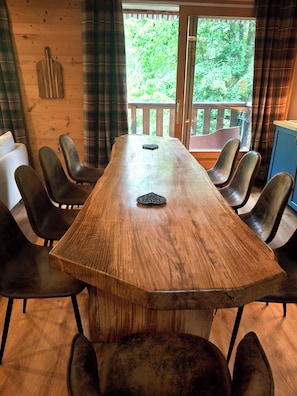 A wooden dining table with eight chairs, a window with a view of greenery, and a plaid curtain. - L'Alpin, an apartment in the heart of the Domaine de la Grande Longe in Oisans (Le Bourg-d'Oisans)
