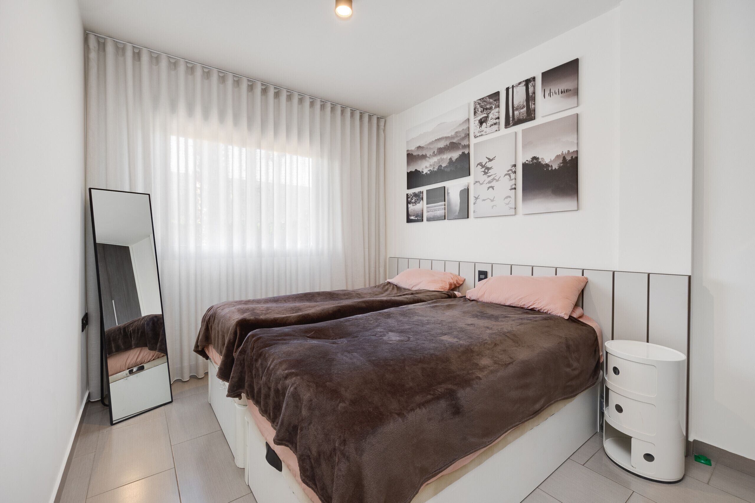 A bedroom with two beds, a mirror, a bedside table, and a wall with framed pictures.
