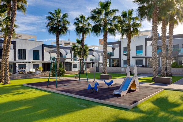 A playground with swings and a slide, surrounded by palm trees and modern buildings. - Relaxing Apartment in La Zenia (Orihuela Costa)
