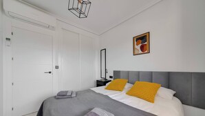 A modern bedroom with a grey upholstered bed, two pillows, a bedside table, a wall-mounted lamp, and a framed abstract painting. - CB Living - Seaview penhouse in Cabo Roig (Orihuela)