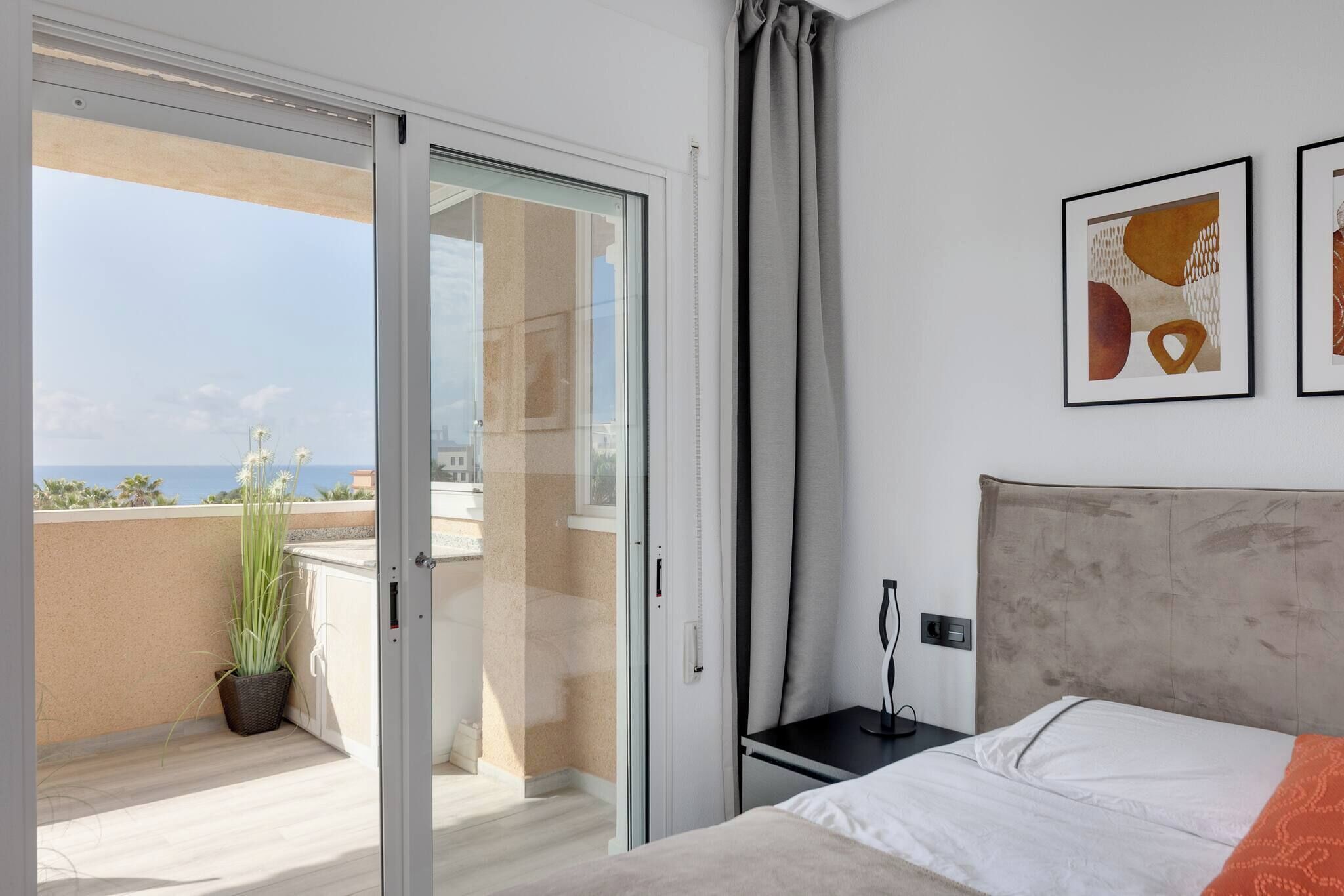 A hotel room with a bed, a nightstand, a potted plant, and a view of the ocean through a sliding glass door.