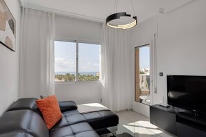 A modern living room with a black leather sofa, a flat-screen TV, and a large window offering a view of palm trees and buildings. - CB Living - Seaview penhouse in Cabo Roig (Orihuela)