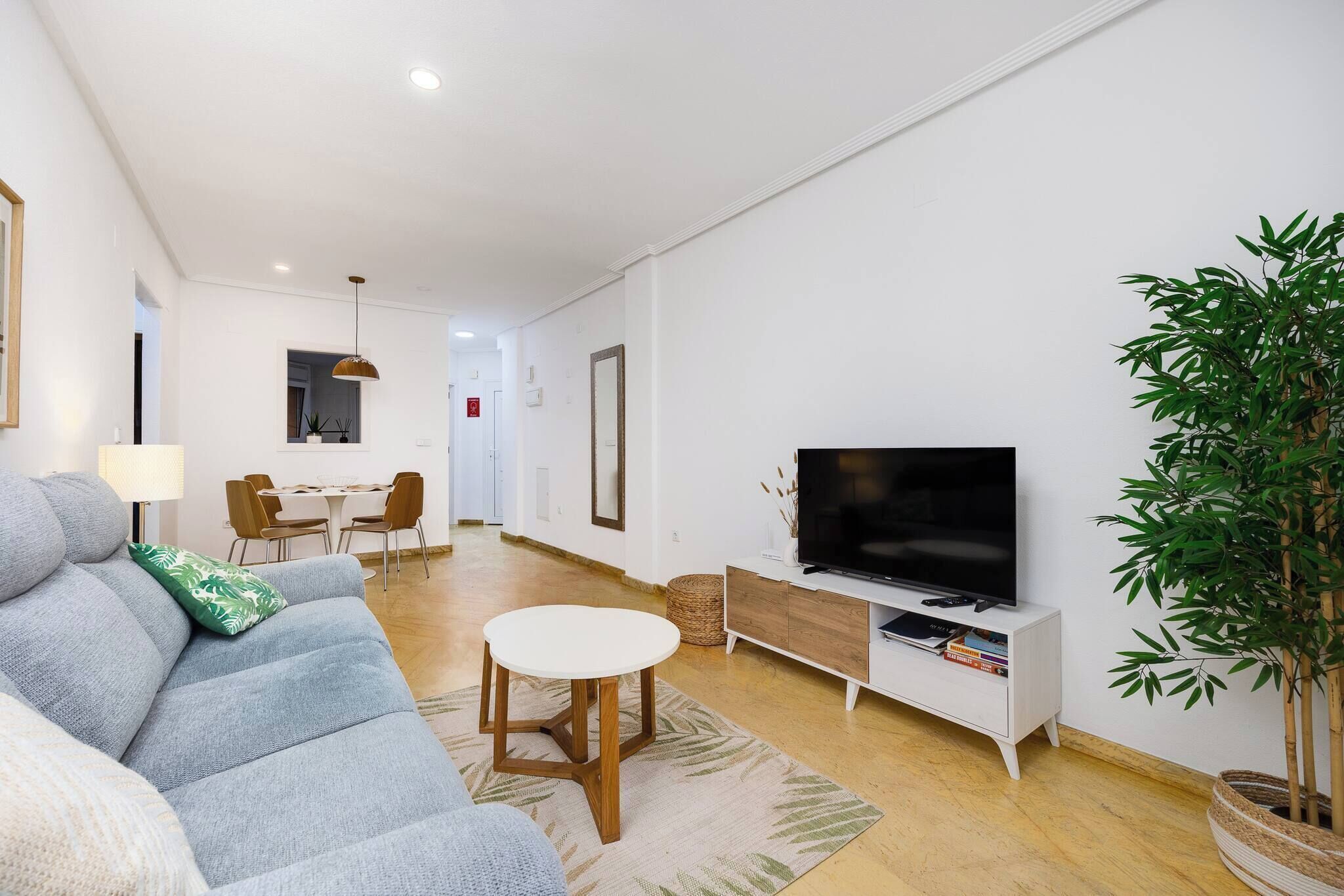 Een moderne woonkamer met een bank, een eettafel, een televisie op een witte kast en een grote plant.