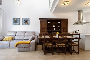 A modern living room with a grey sofa, wooden dining table, and chairs. There is a cabinet with shelves and a kitchen area with a stove. - CB Living - Cosy loft with terrace, Villamartin (Orihuela)
