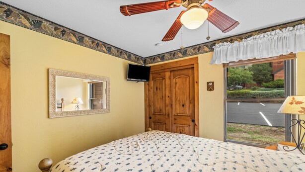 Un dormitorio con una cama, ventilador de techo, televisión, ventana con vista a la calle y una puerta.