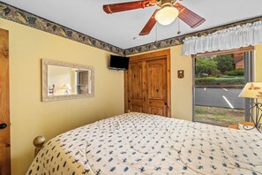 Une chambre à coucher comprenant un lit, un ventilateur de plafond, une télévision, une fenêtre donnant sur la rue et une porte.