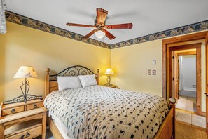 Quarto com cama, duas mesas de cabeceira com luminárias, um ventilador de teto e uma porta para o banheiro.
