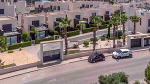 A residential area with modern buildings, a road with cars, and a clear view of the sea in the background. - CB Living - 3 bedroom home in Punta Prima (Alicante / Alacant)