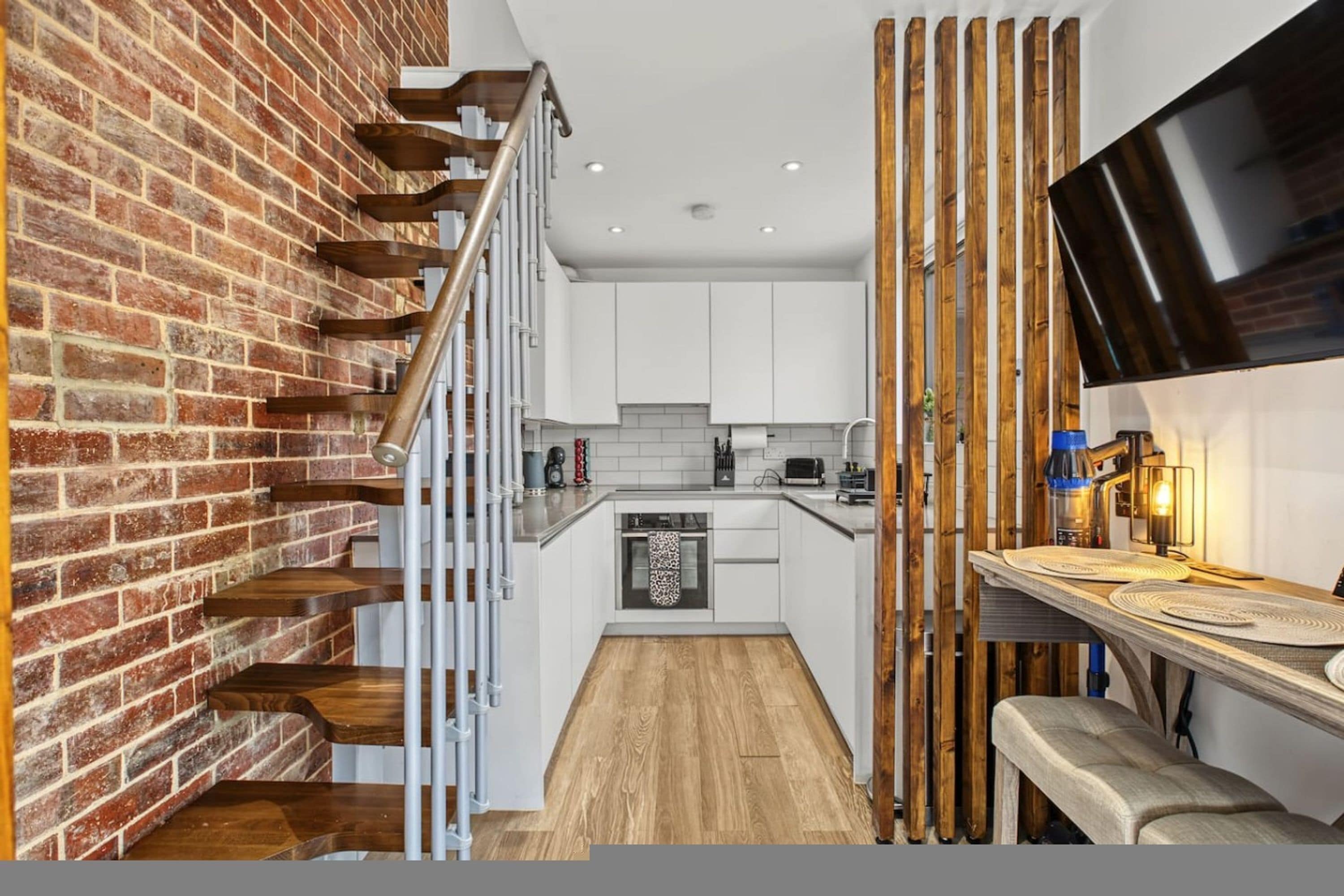 A kitchen with white cabinets, a wooden staircase, and a brick wall.