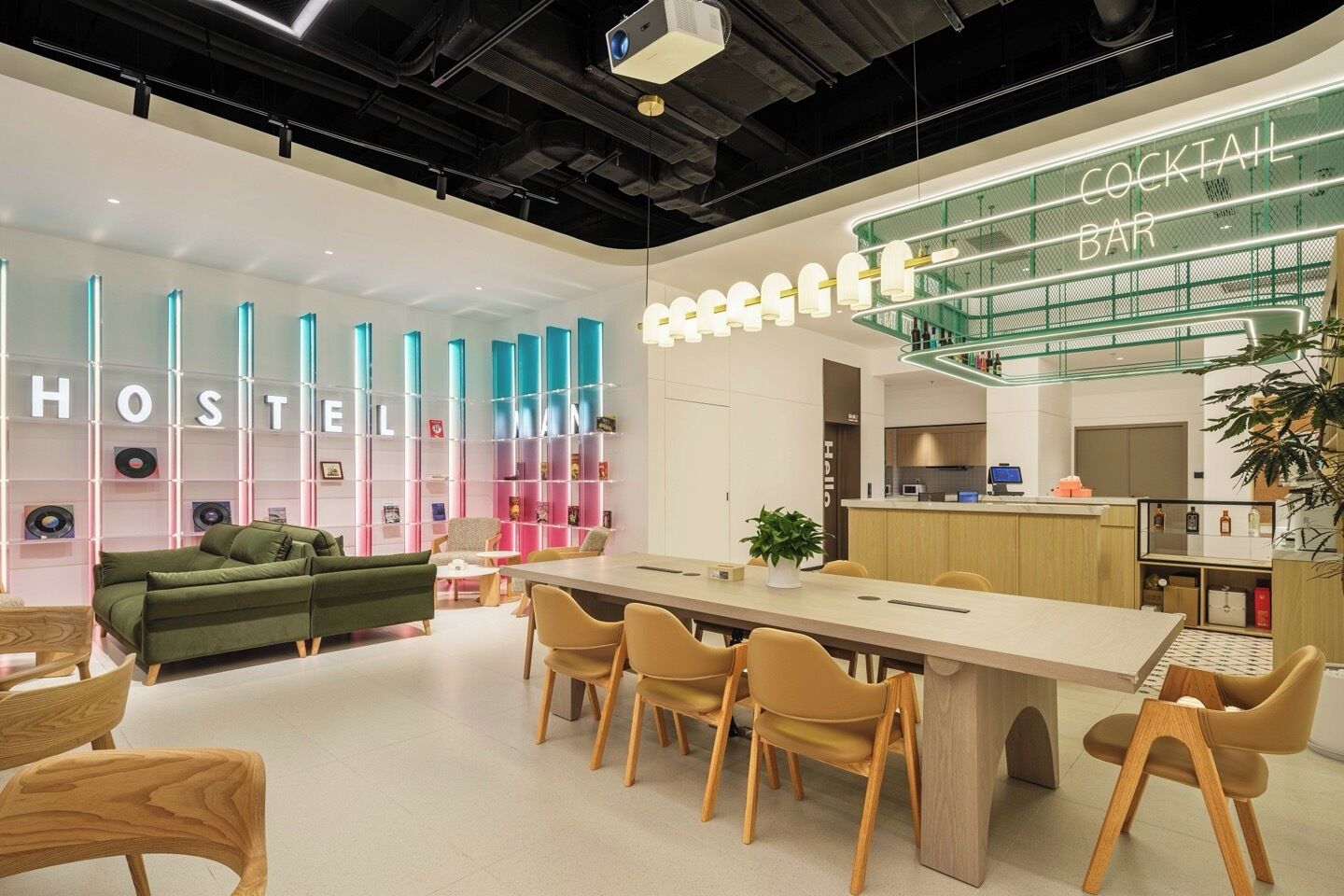 A modern hostel interior with a cocktail bar, a communal dining area, and a bookshelf with colorful lighting.