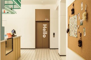 Un espace d’accueil d’hôtel comprenant un comptoir de réception, une pancarte « Bonjour » sur la porte, un mur orné de photos de voyage et un sol à motifs.
