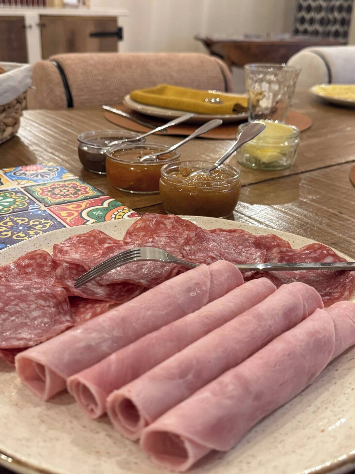 A table with a plate of sliced salami, a jar of jam, and a glass of water.