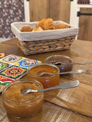 A wooden table with three jars of jam, a spoon, and a basket of bread.