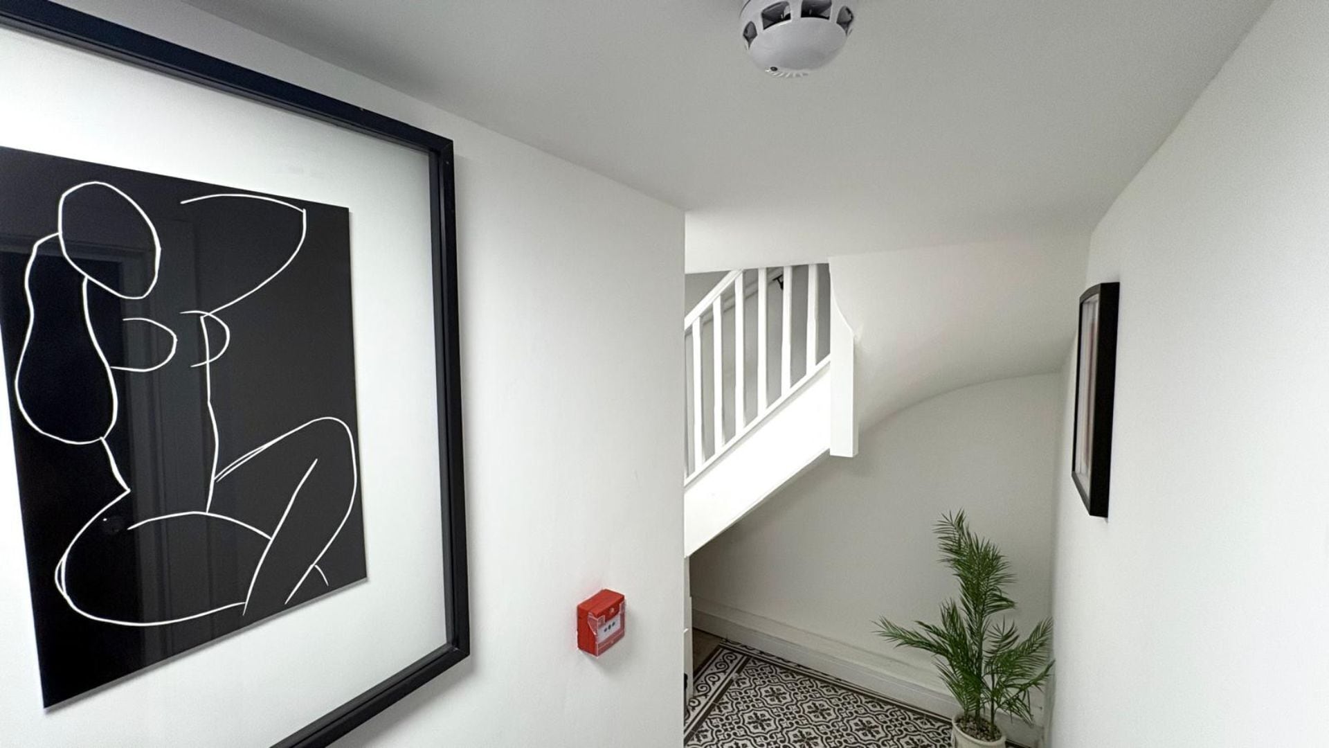 A hallway with a framed abstract artwork, a staircase with white railings, a potted plant, and a patterned floor.