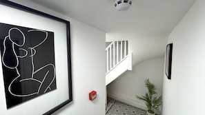 Un couloir orné d’une œuvre d’art abstraite encadrée, un escalier avec des rampes blanches, une plante en pot et un sol à motifs.
