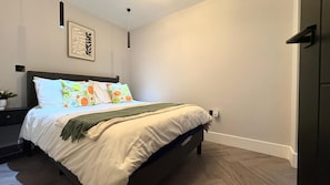 Une chambre  à coucher avec un lit, une table de chevet, une lampe fixée au mur et un tableau encadré.