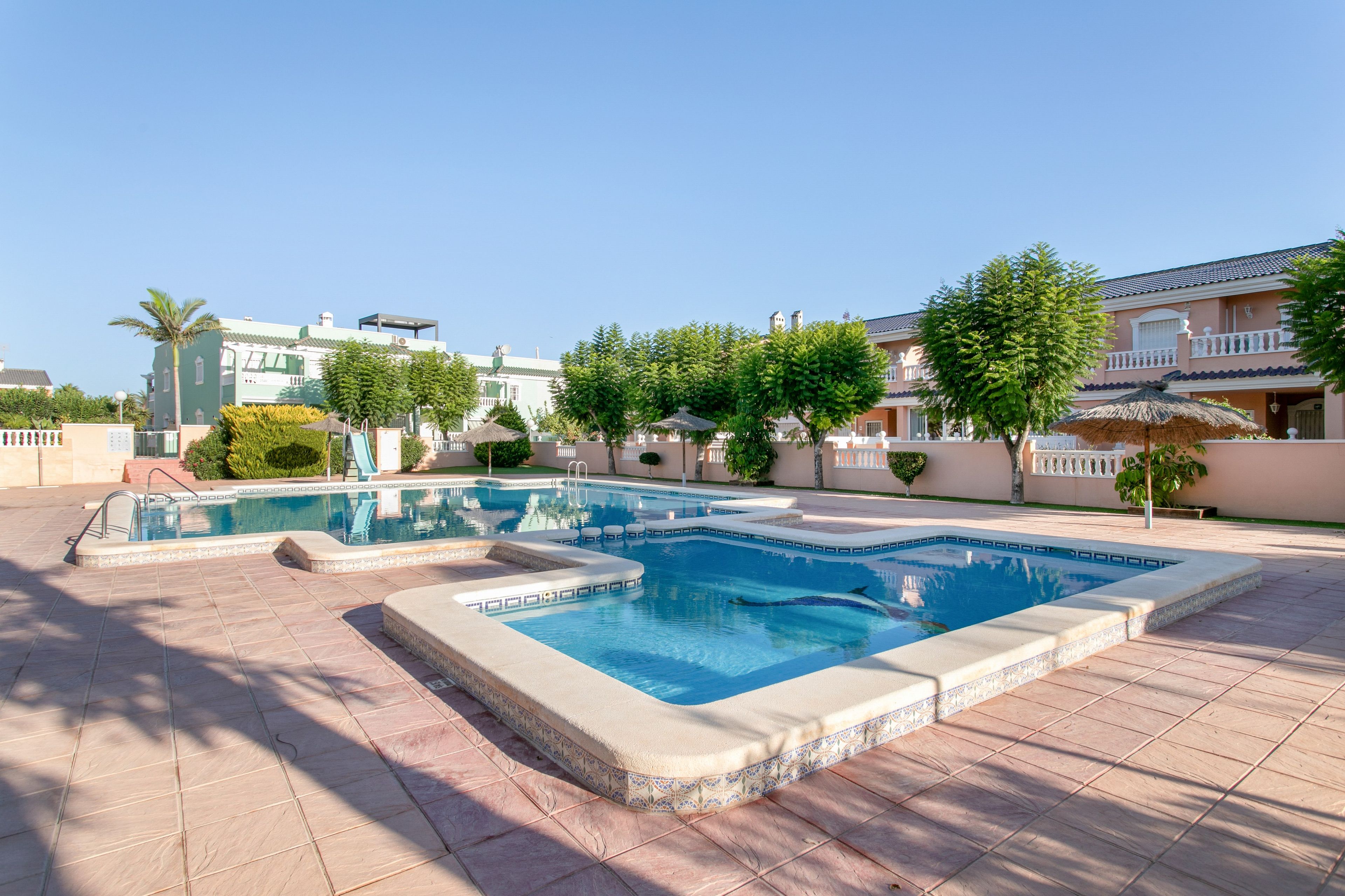 Une piscine dotée d’une terrasse carrelée, entourée de verdure et de bâtiments.