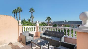A balcony with wicker furniture, a tiled floor, and a view of palm trees and buildings.