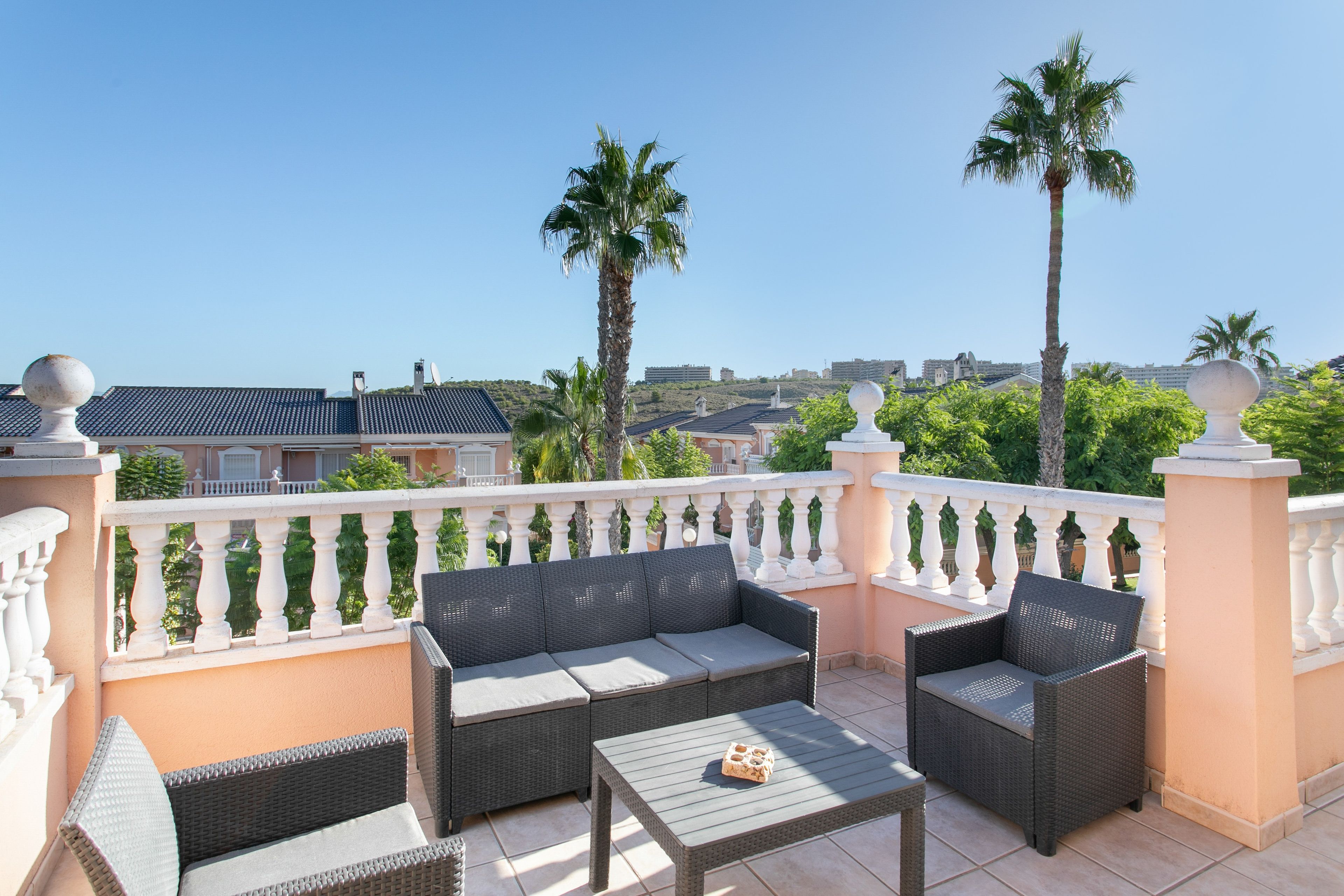 Een balkon met rieten meubels, een tafeltje en uitzicht op gebouwen en palmbomen.