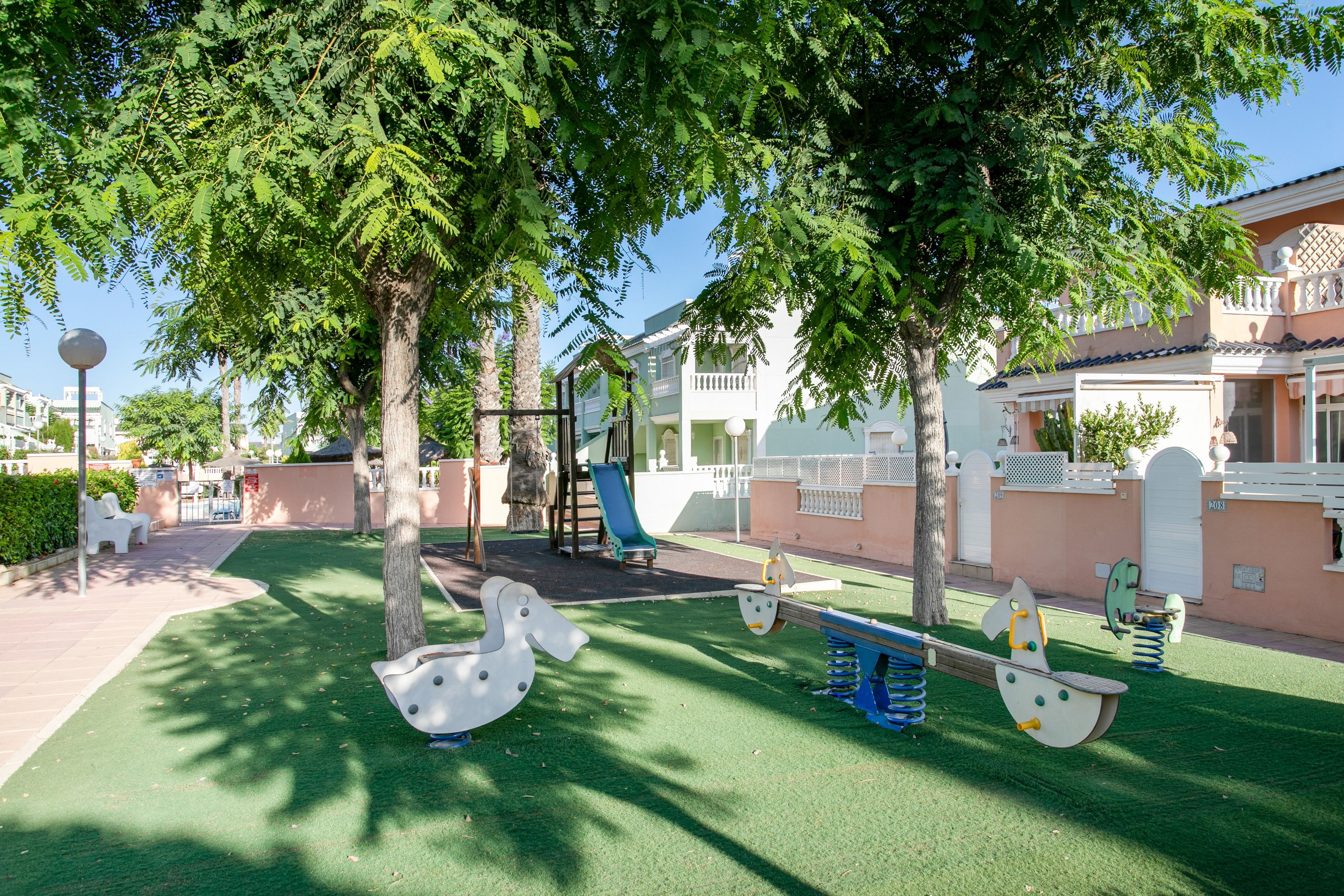 A playground with swings and a slide, surrounded by trees and a building with a balcony.