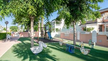 A playground with swings and a slide, surrounded by trees and a building with a balcony.