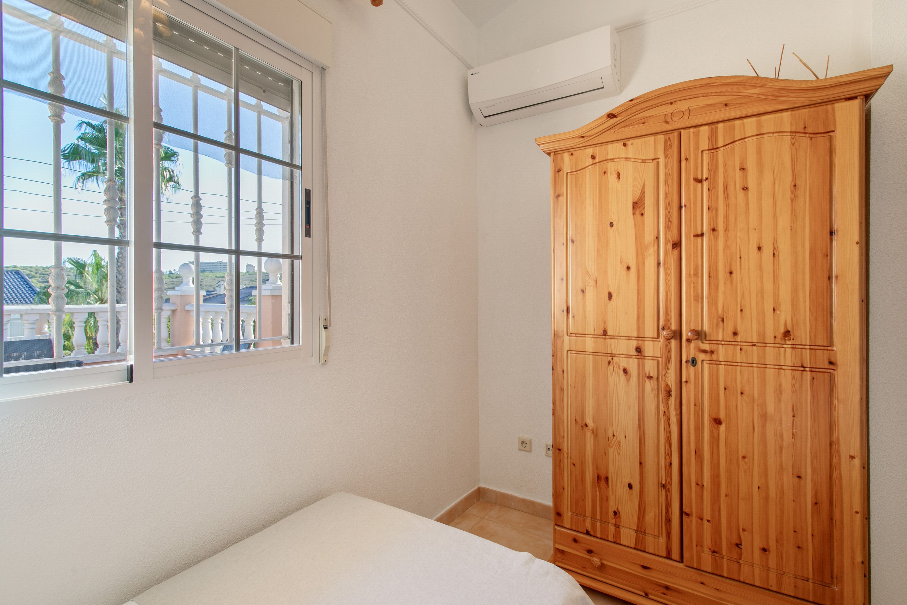A bedroom with a large wooden wardrobe, a single bed, a window with a view of palm trees, and an air conditioning unit on the wall.