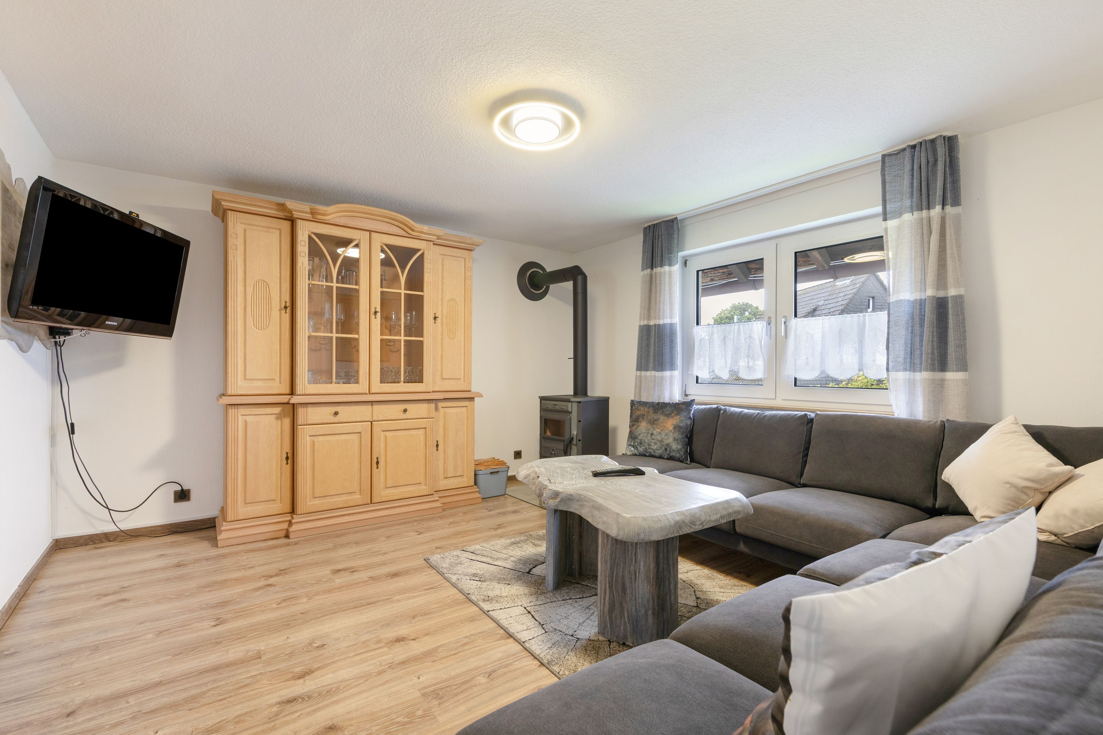 En stue med et træskab, en sofa, et fjernsyn ophængt på væggen og et vindue med gardiner.