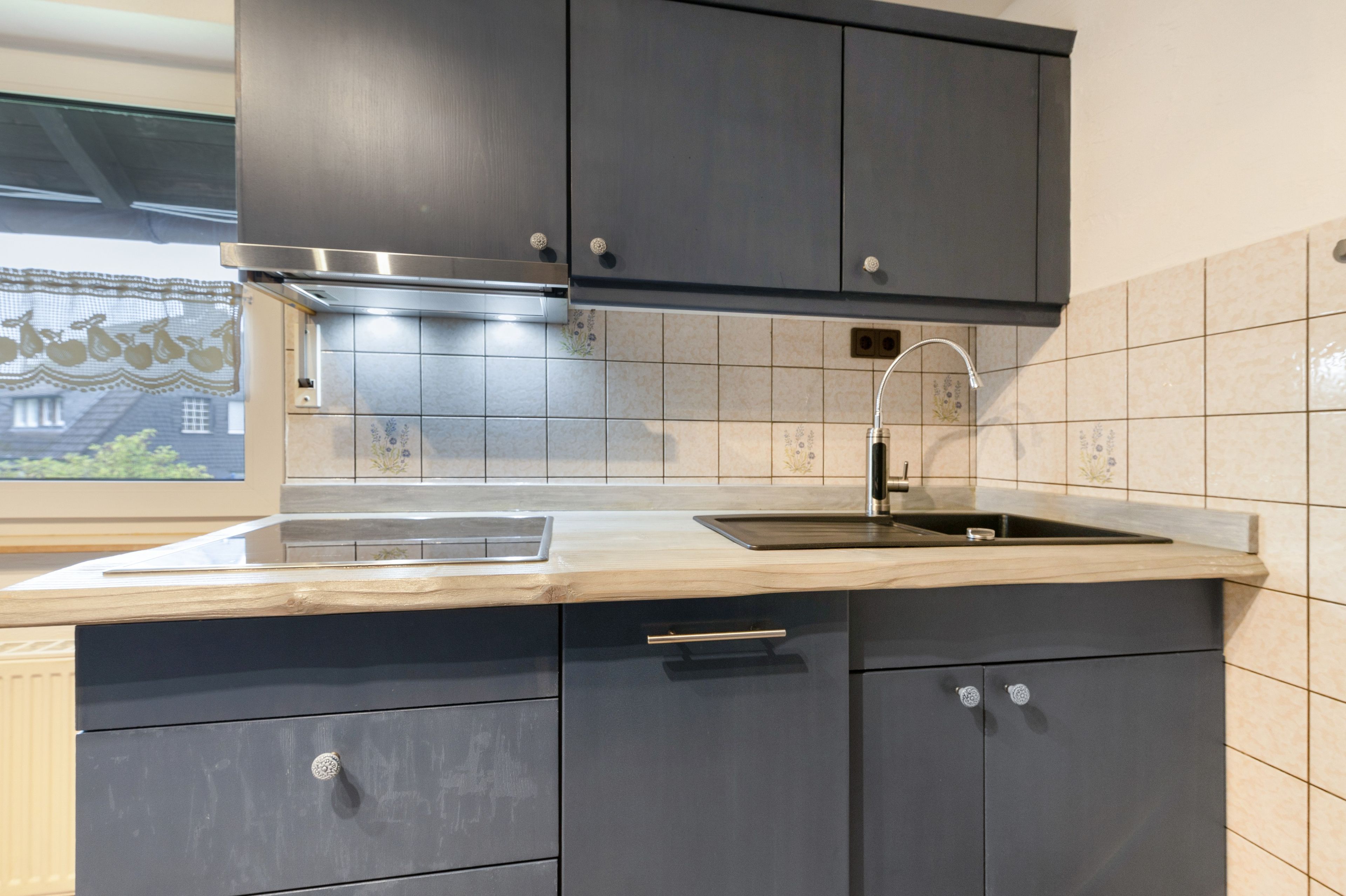 A modern kitchen with dark cabinetry, a stainless steel range hood, a sink, and a window with a view of a building.