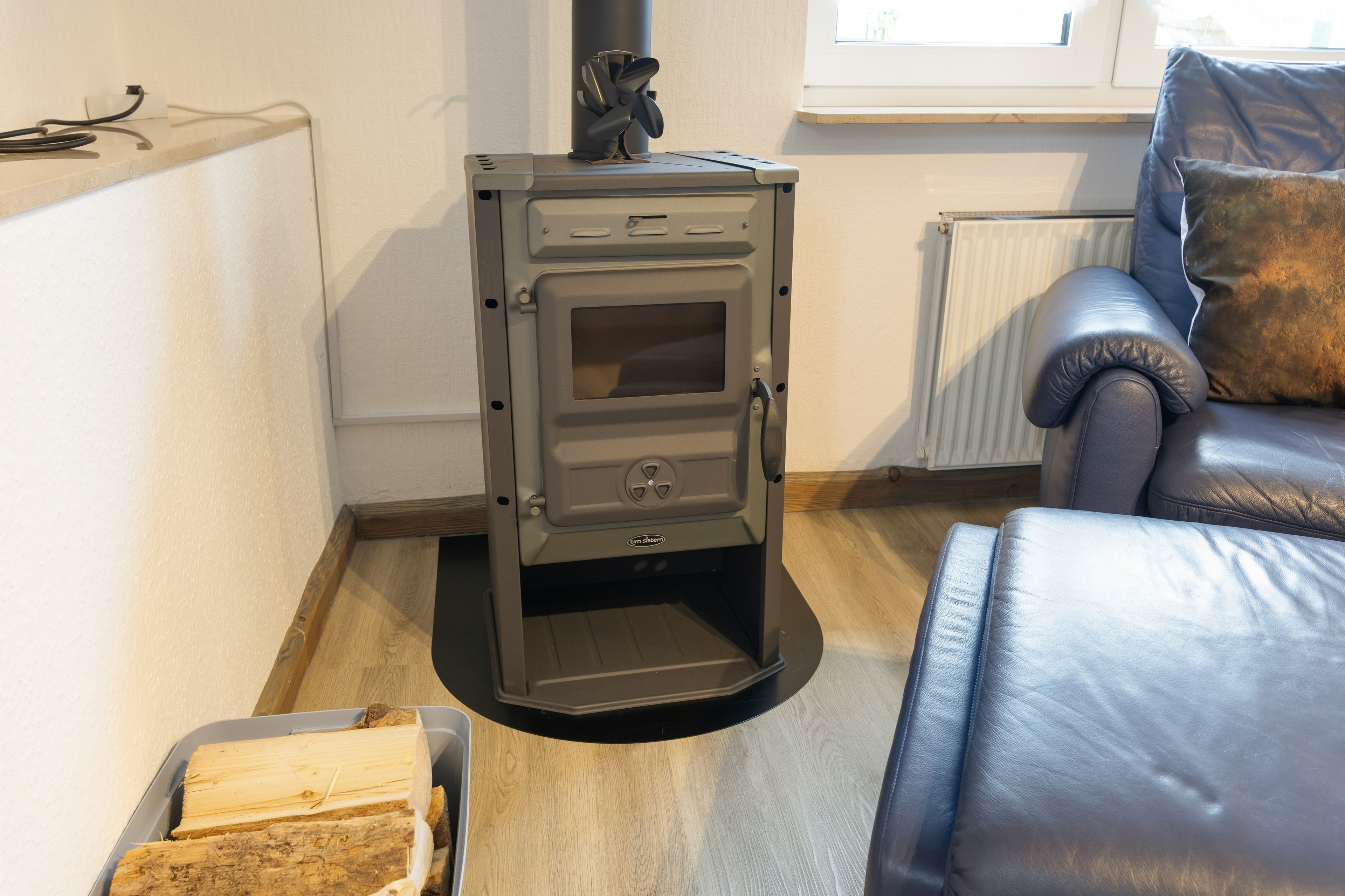 Ein kleiner Holzofen mit Bedienfeld, ein Fenster darüber und ein Ledersofa im Raum.