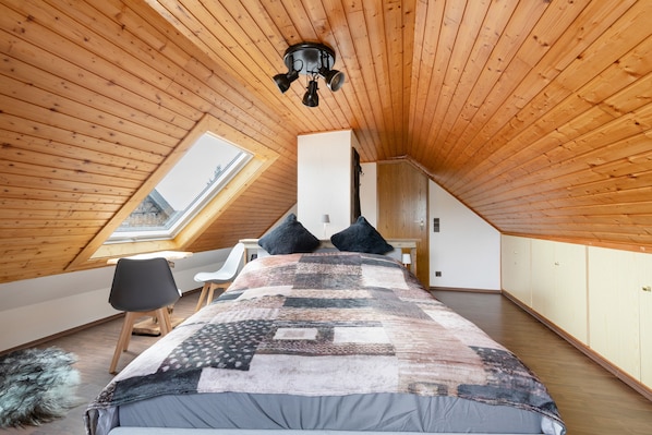 A high-ceilinged room with a sloped wooden ceiling, a skylight, a bed with a patterned blanket, a desk with a chair, and a door.
