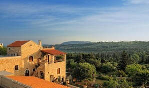 A stone building with a red-tiled roof, a balcony, and a view of a forested hillside.