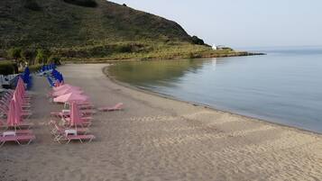 Une plage bordée de rangées de chaises longues et de parasols, une mer calme et une colline en arrière-plan.
