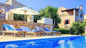 A poolside area with lounge chairs and an umbrella, surrounded by stone walls and vegetation.