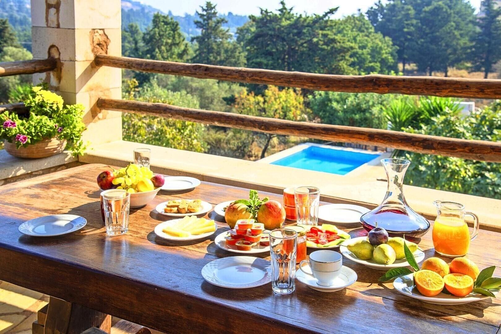 Ein Esstisch mit verschiedenen Früchten, Gläsern und einer Saftkanne, mit Blick auf einen Swimmingpool und Grünflächen.