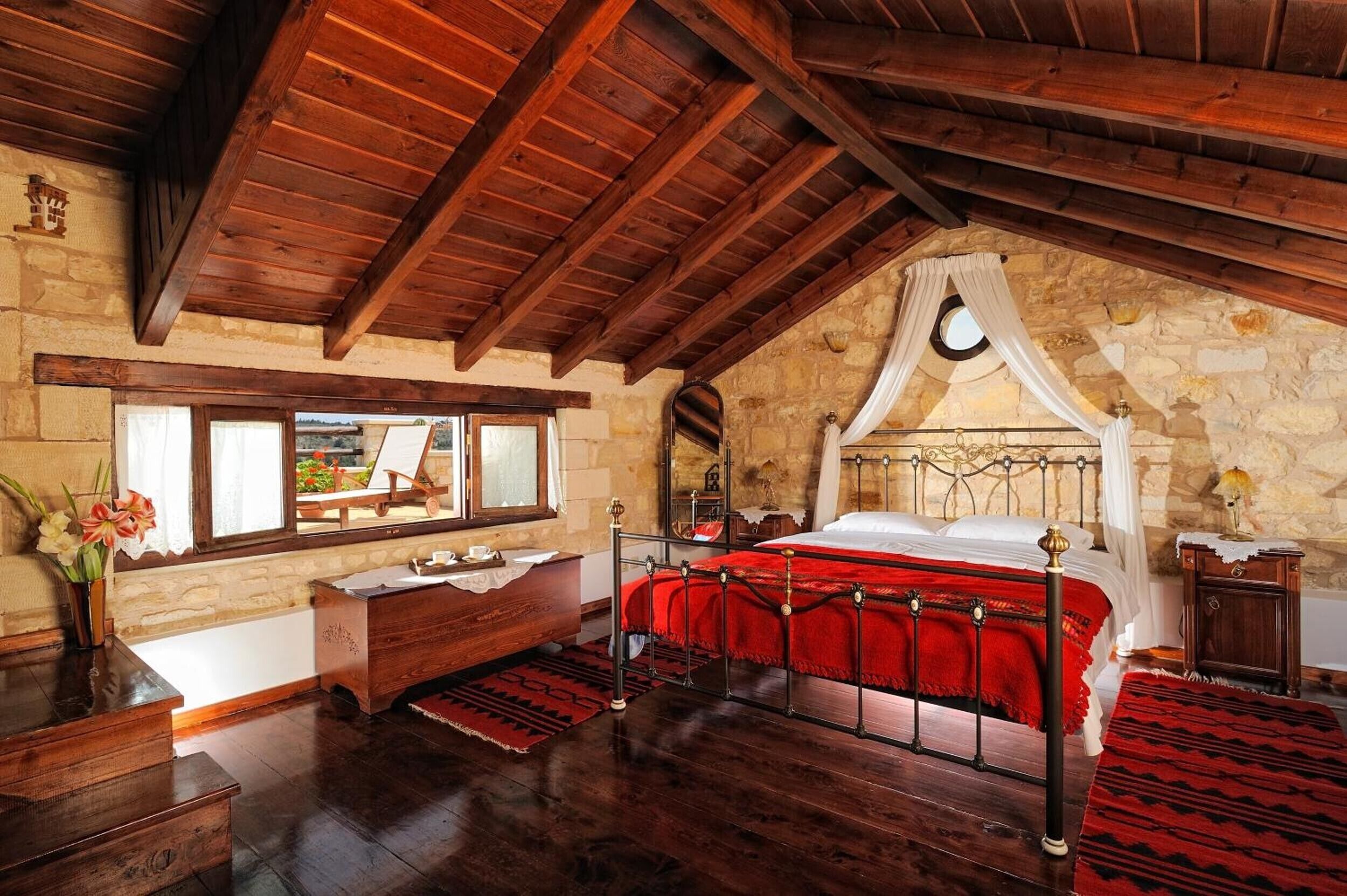 A room with a wooden ceiling, stone walls, a bed with a red bedspread, a wooden dresser, and a window with a view.