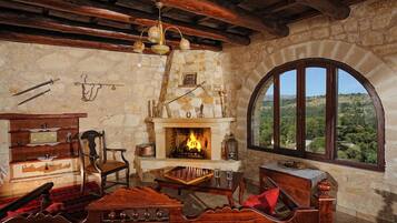A rustic room with a stone fireplace, wooden furniture, and a view through a large arched window.