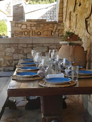 Une table à manger dressée avec de l’assiette bleue, des verres transparents et des couverts, avec un mur de pierre et une enseigne indiquant « Strofilia Villas » en arrière-plan.