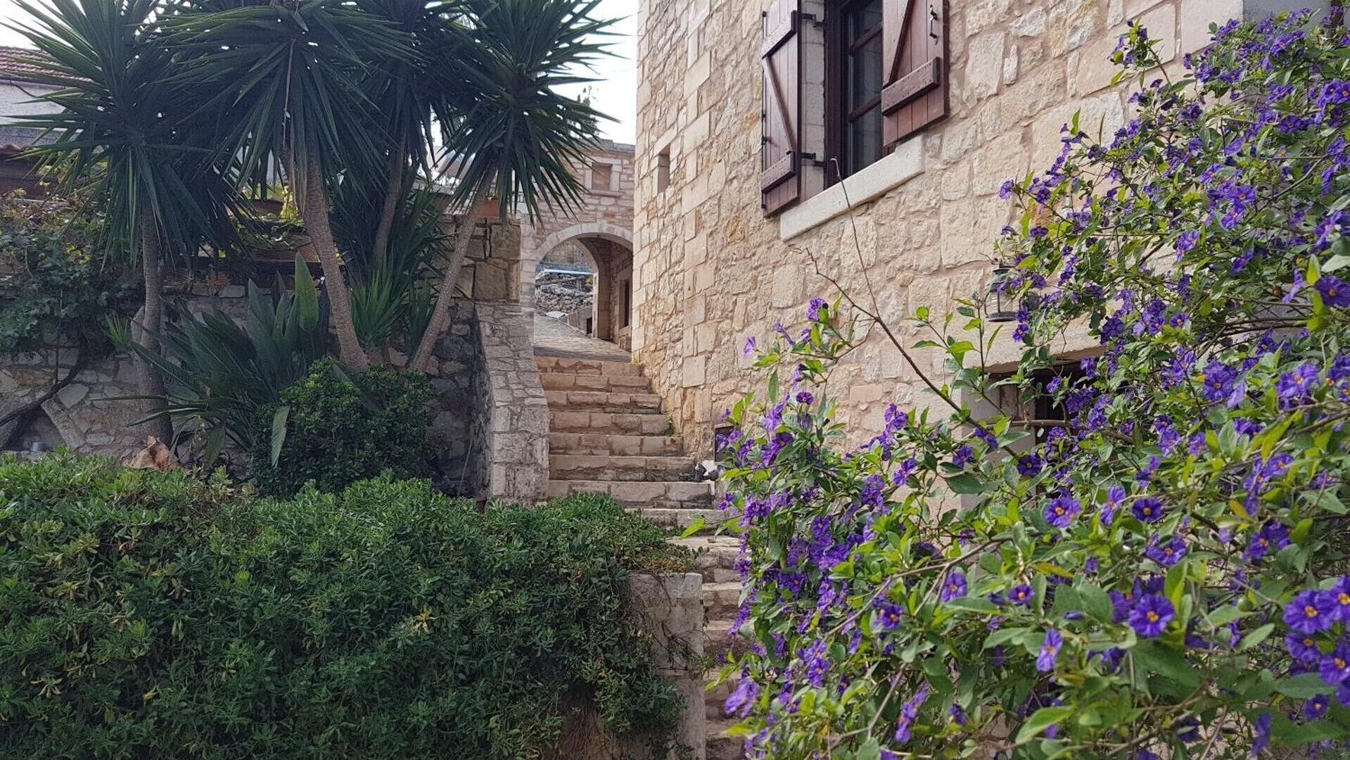 A stone building with a staircase leading to an archway, surrounded by lush greenery and purple flowers.