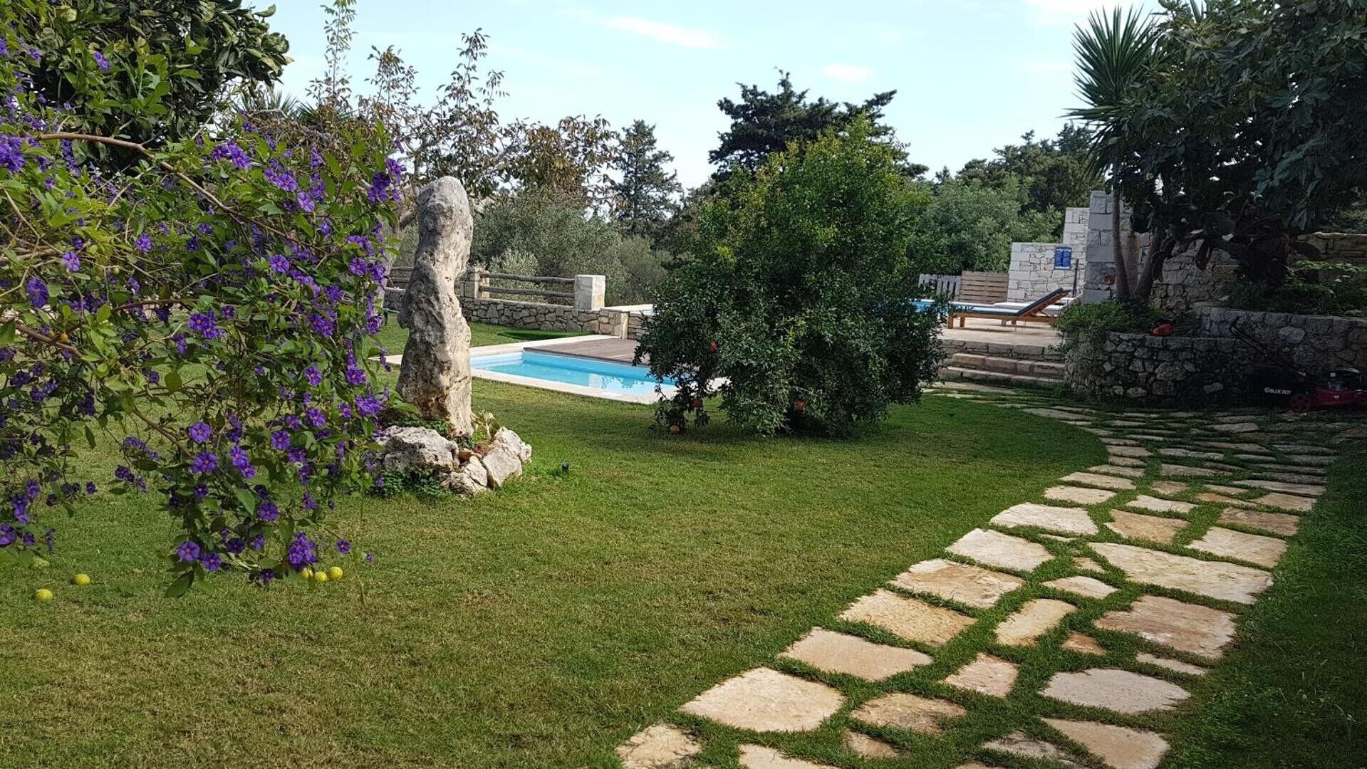 Un jardin avec une piscine, un chemin de pierre et une variété de plantes, notamment des fleurs violettes.