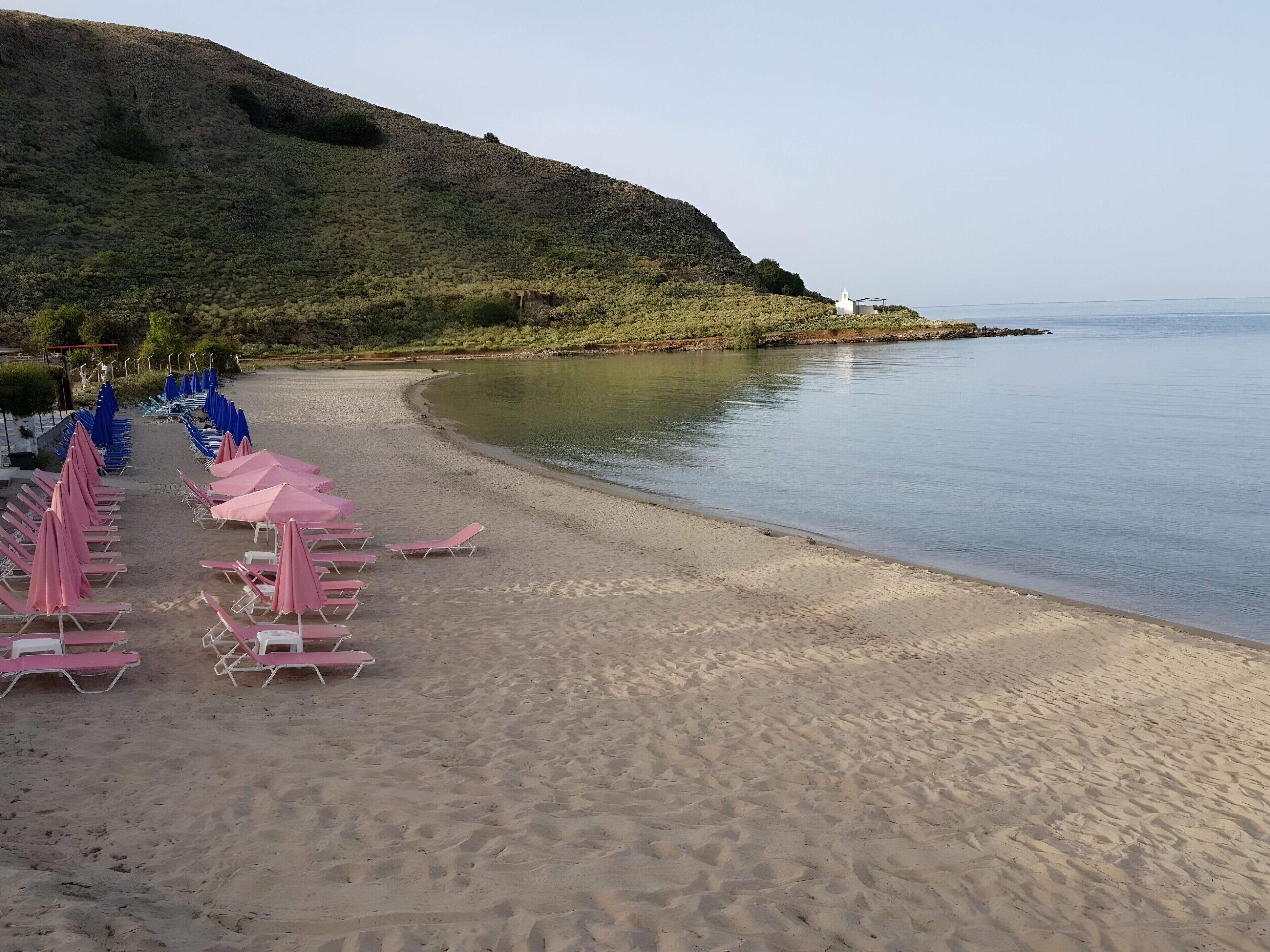 A beach with rows of sun loungers and umbrellas, a calm sea, and a hill in the background.