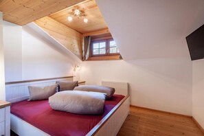 Ein Schlafzimmer mit schrägen Wänden, einem fensterförmigen Öffnung mit Vorhängen, einem Bett mit Kissen und einer Heizung.