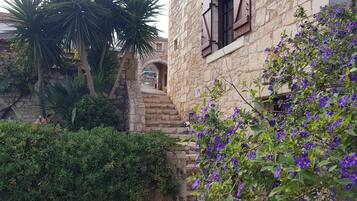 Un edificio in pietra con una scalinata che conduce a un arco, immerso nel verde e circondato da fiori viola.