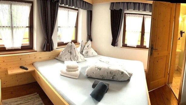 Um quarto com uma cama, uma escrivaninha de madeira, uma janela com cortinas, uma porta de madeira, um sofá e uma televisão fixada na parede.