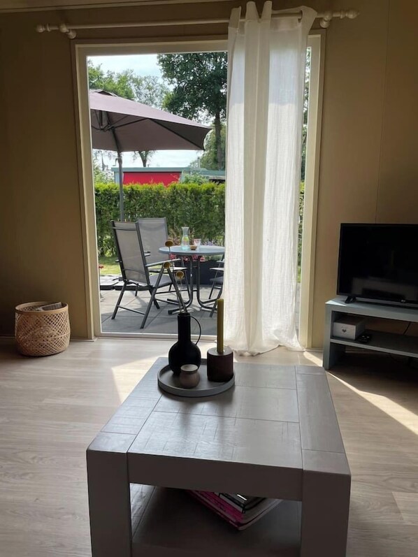 A room with a television, a table with a vase, and a view of an outdoor patio with chairs and a table.