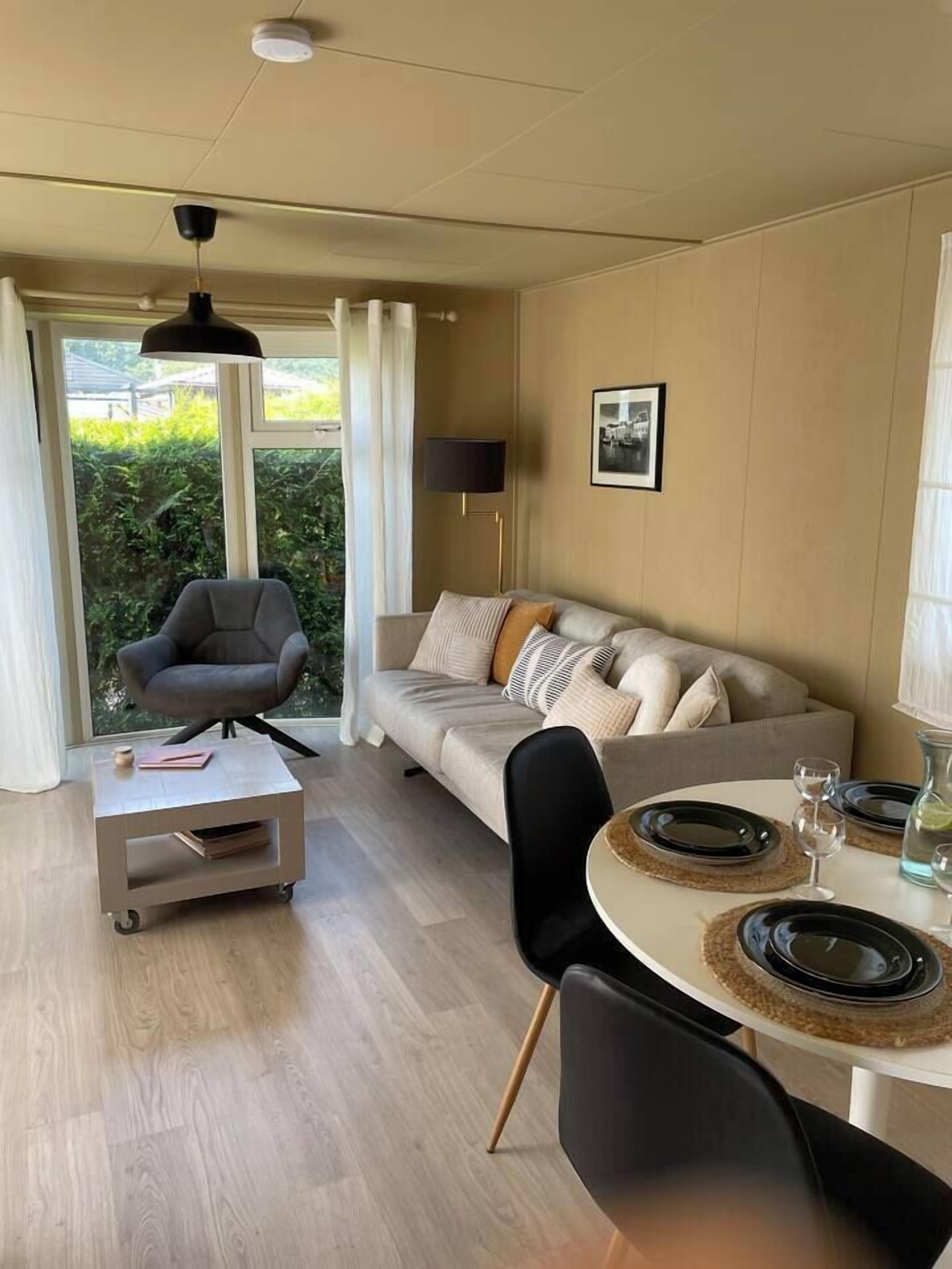 A modern living room with a sofa, a dining table set with plates and glasses, a chair, and a small table with a lamp.