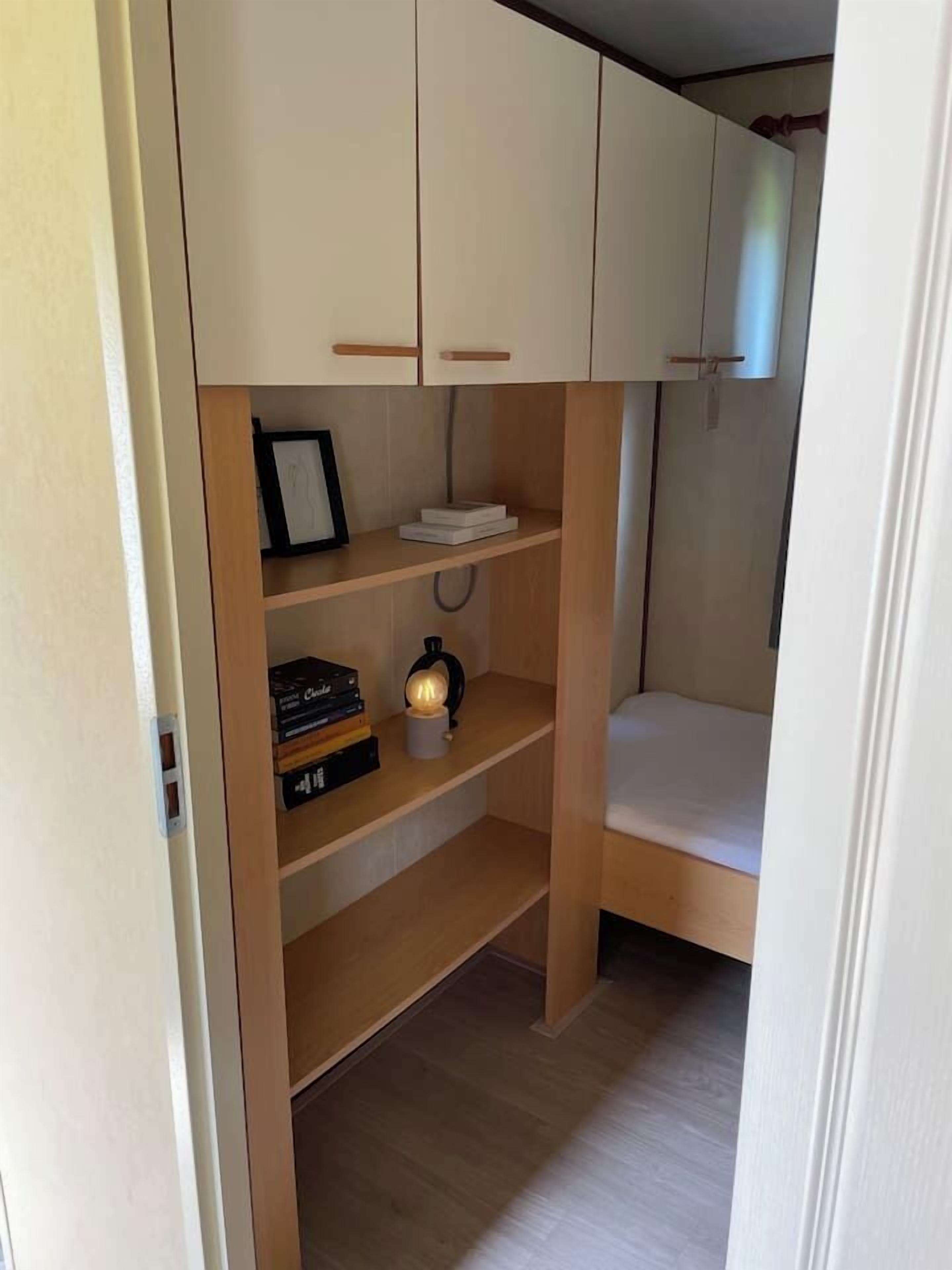 Een kleine kamer met een bed, een plank vol boeken en een lamp, en een klein bureau.