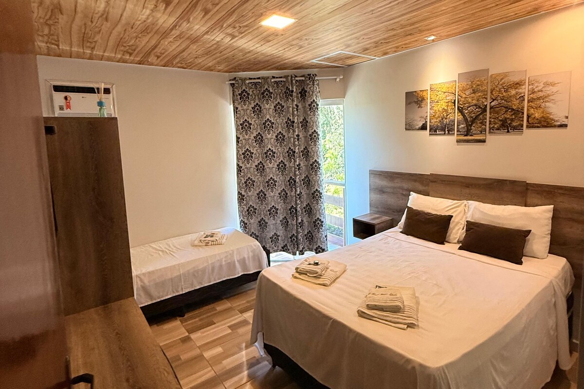 A hotel room with a wooden ceiling, two beds, a nightstand, a wall-mounted air conditioning unit, and a window with curtains.