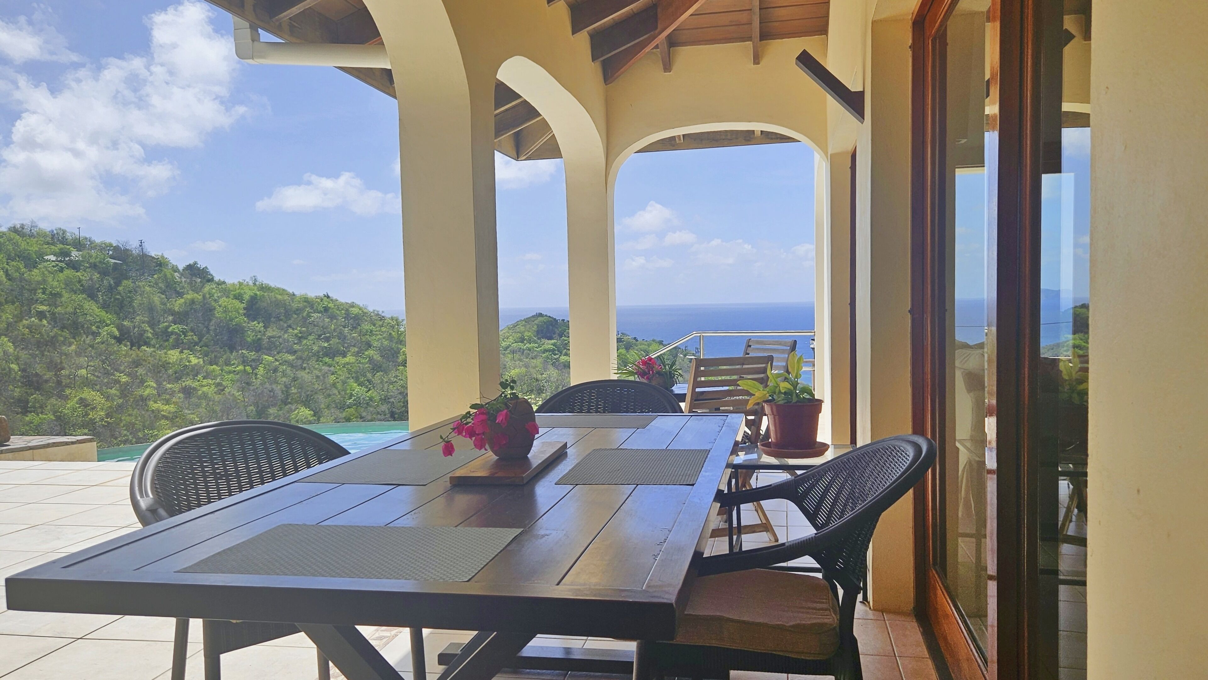 A covered outdoor seating area with a table and chairs, overlooking a scenic view of the ocean and hills.
